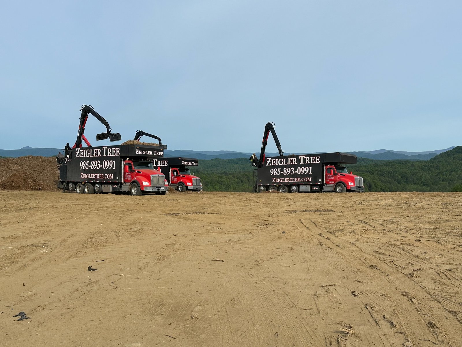 Two grapple trucks loading debris at a land-clearing site